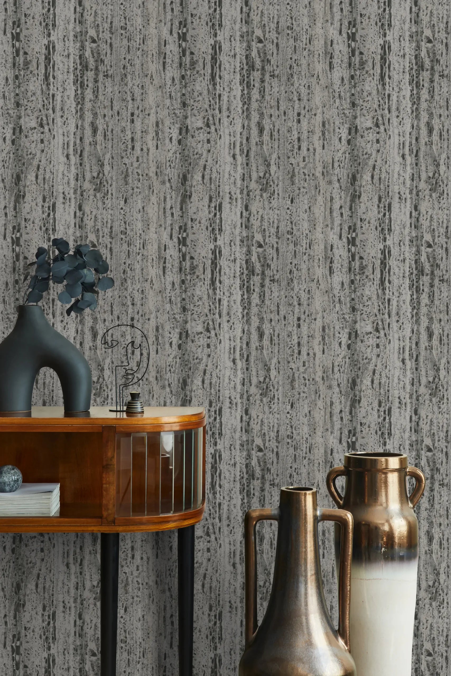 Marburg Travertino 33354 Gray stone-effect wallpaper with vertical travertine-style streaks, shown behind a wood console with a matte sculptural vase and foliage, plus two tall metallic handled vases.