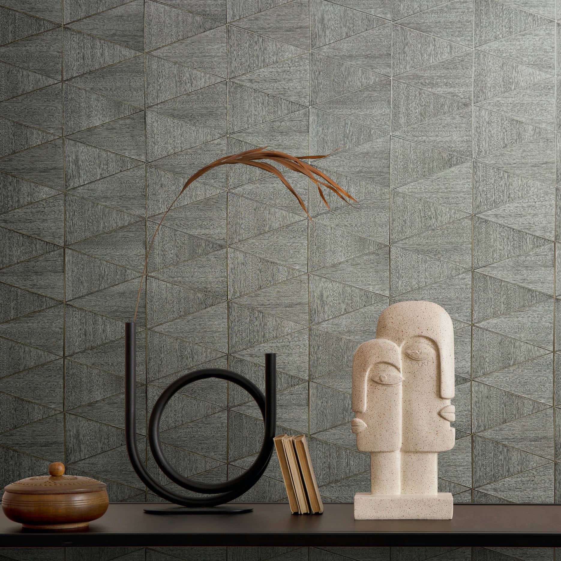 Marburg Travertino 33355 Gray geometric triangle wallpaper shown behind a console with a black circular sculpture holding a dried stem, an abstract stone bust, a few books, and a small lidded wooden bowl.
