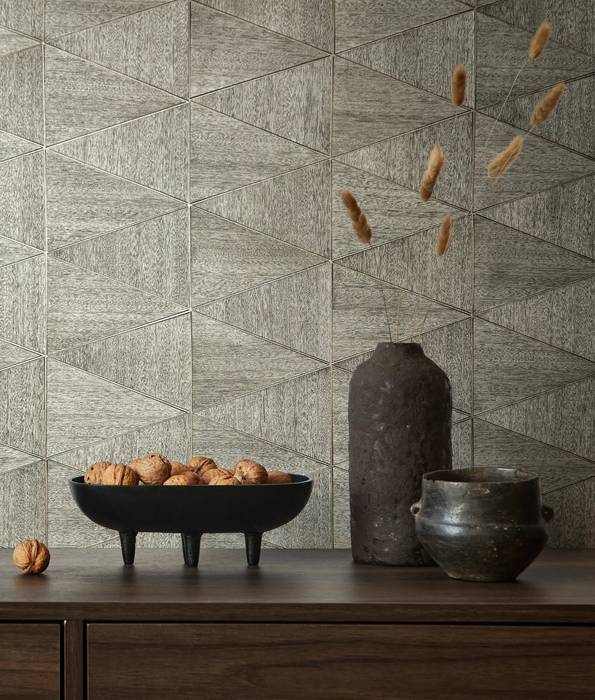 Marburg Travertino 33355 Gray geometric triangle-patterned wallpaper behind a wood console holding a black footed bowl of walnuts, a tall dark vase with dried stems, and a small dark pot.
