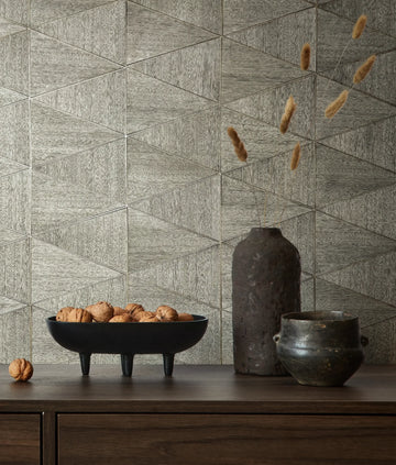 Marburg Travertino 33355 Gray geometric triangle-patterned wallpaper behind a wood console holding a black footed bowl of walnuts, a tall dark vase with dried stems, and a small dark pot.