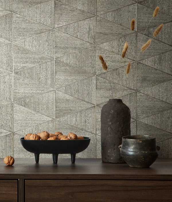 Marburg Travertino 33355 Gray geometric triangle-patterned wallpaper behind a wood console holding a black footed bowl of walnuts, a tall dark vase with dried stems, and a small dark pot.