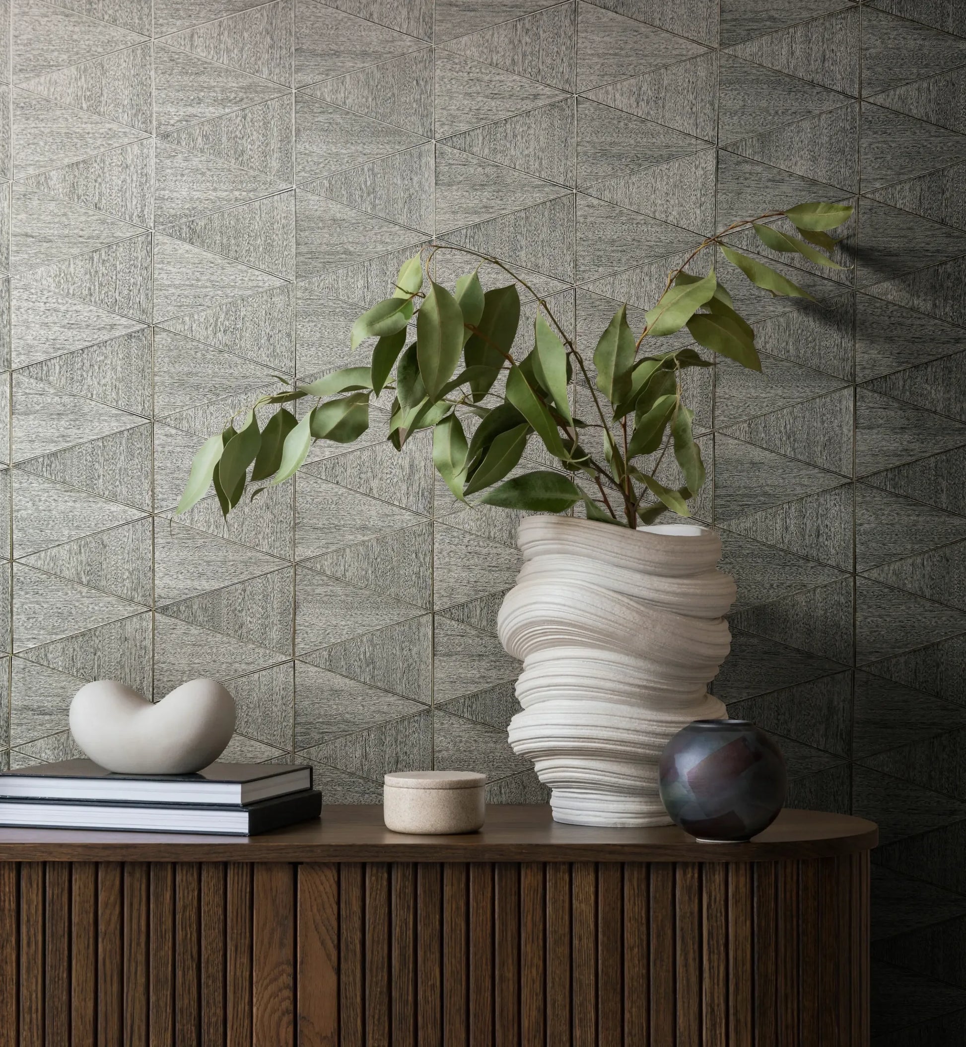 Marburg Travertino 33355 Gray triangle-pattern wallpaper behind a wood console styled with a sculptural white vase holding green leaves, stacked books with a small decorative object, a small round container, and a dark ceramic vessel.