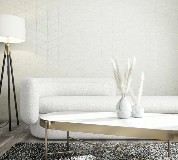 Marburg Travertino 33356 White wallpaper with a subtle geometric triangle line pattern behind a white sofa, a black tripod floor lamp, and a brass-framed coffee table with two small vases of pampas stems.