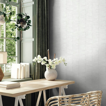 Marburg Travertino 33356 Light gray-white geometric wallpaper with a subtle faceted triangle pattern shown beside a windowed workspace with a wooden desk, woven chair, green curtains, hanging plant, books, and a vase of white flowers.