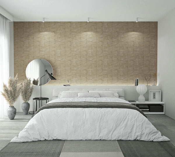Marburg Travertino 33359 Bedroom scene featuring a beige geometric textured wallpaper feature wall behind a low bed with white bedding, a round mirror and black side table with lamp on the left, floor vases with pampas grass, and a white nightstand with a ribbed lamp on the right.
