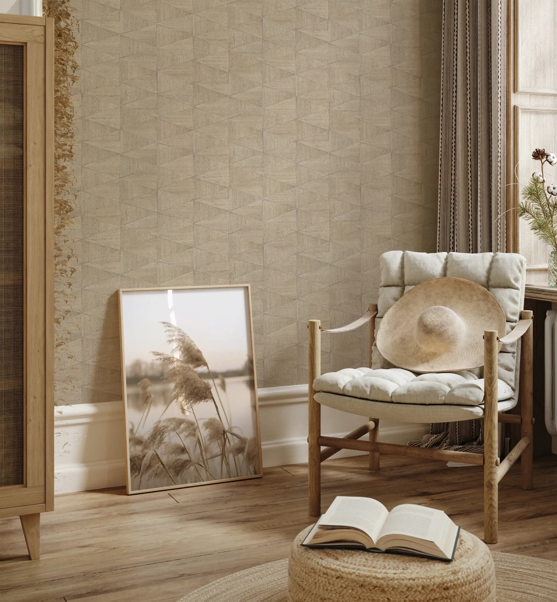 Marburg Travertino 33359 Beige and brown geometric wallpaper with a small triangular pattern behind a cushioned wooden chair holding a straw hat. A framed photo leans against the wall beside a woven pouf with an open book, near a window with curtains.