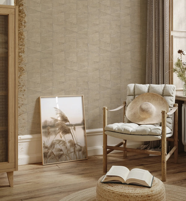 Marburg Travertino 33359 Beige and brown geometric wallpaper with a small triangular pattern behind a cushioned wooden chair holding a straw hat. A framed photo leans against the wall beside a woven pouf with an open book, near a window with curtains.