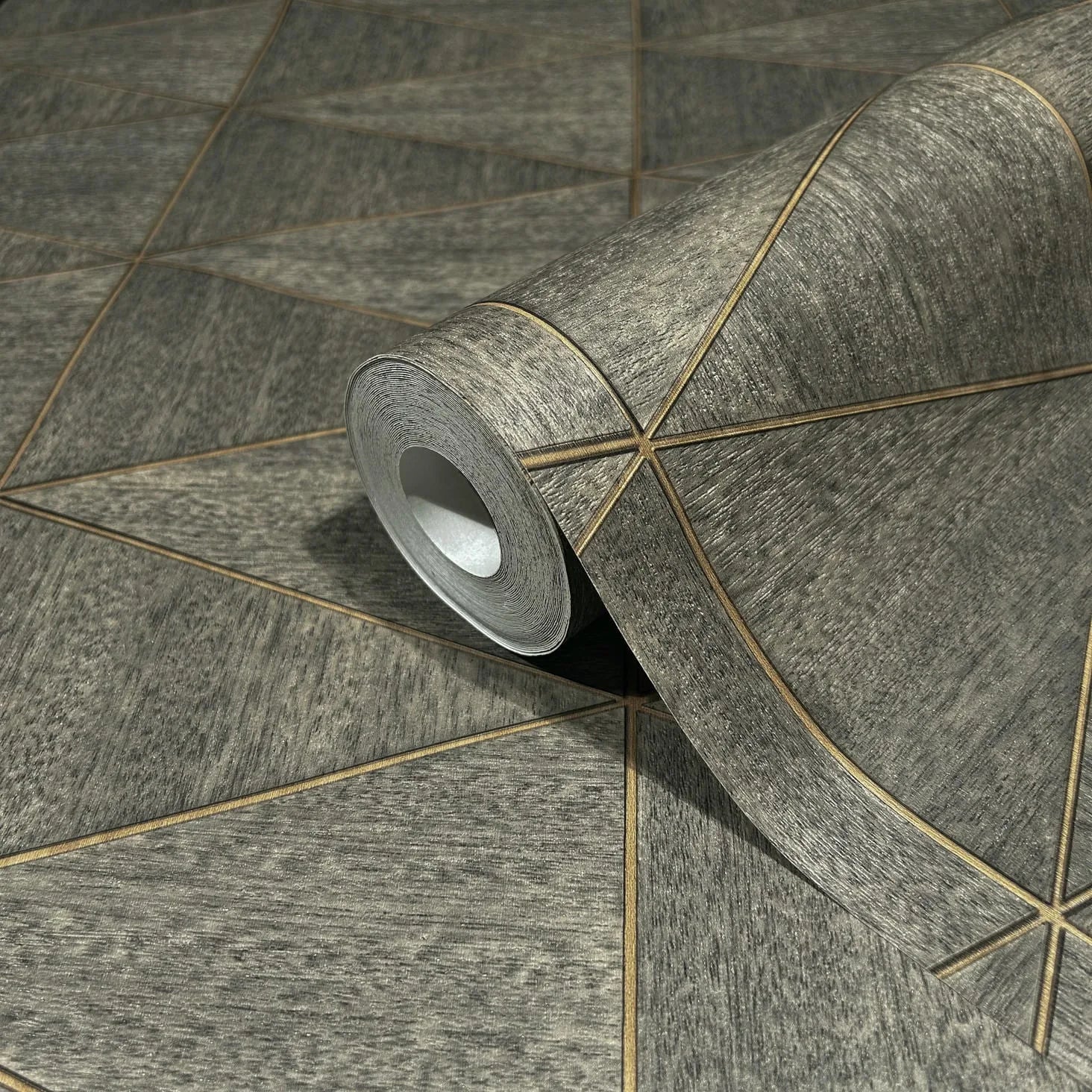 Marburg Travertino 33360 A wallpaper roll resting on the same pattern, showing a textured brown-gray surface crossed by thin gold-toned lines that create large triangular shapes.