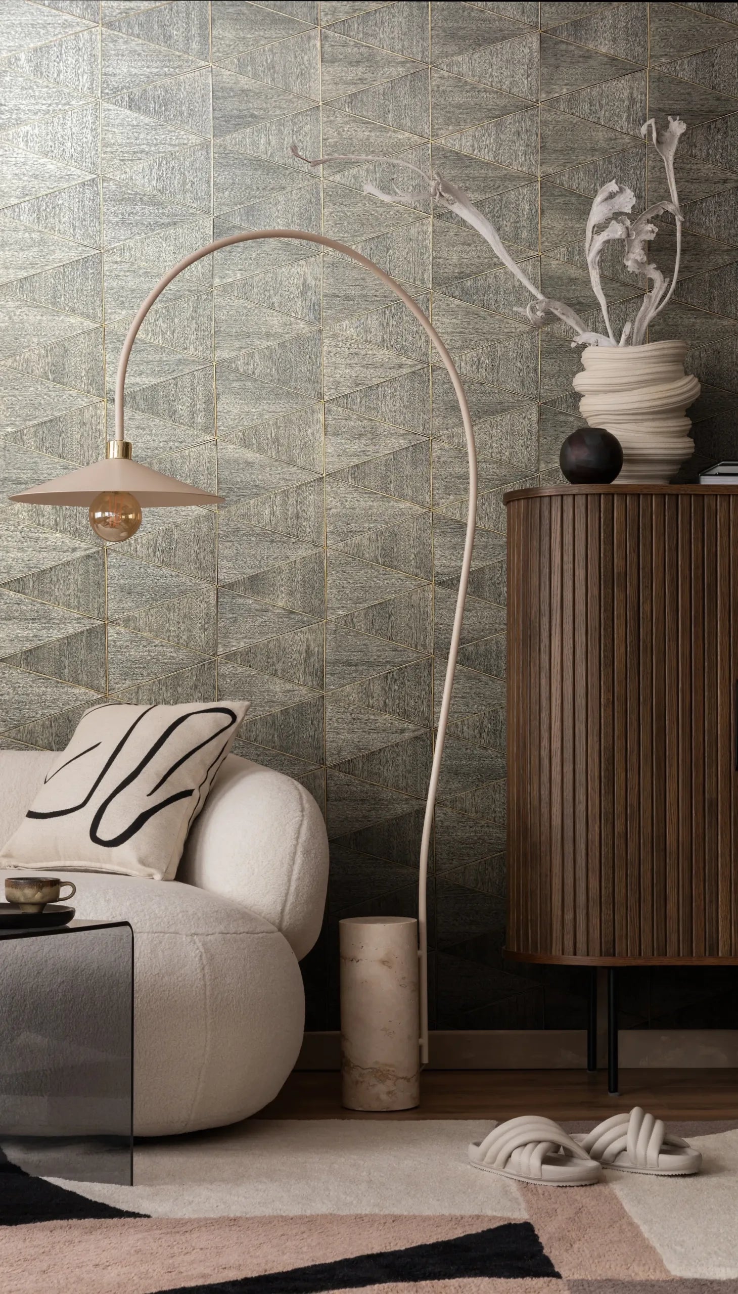Marburg Travertino 33360 Geometric triangle-patterned wallpaper with a stone-like texture and thin gold lines, shown in a living room with a curved white lounge chair, arc floor lamp, fluted wood cabinet, glass side table, and sculptural vase with branches.