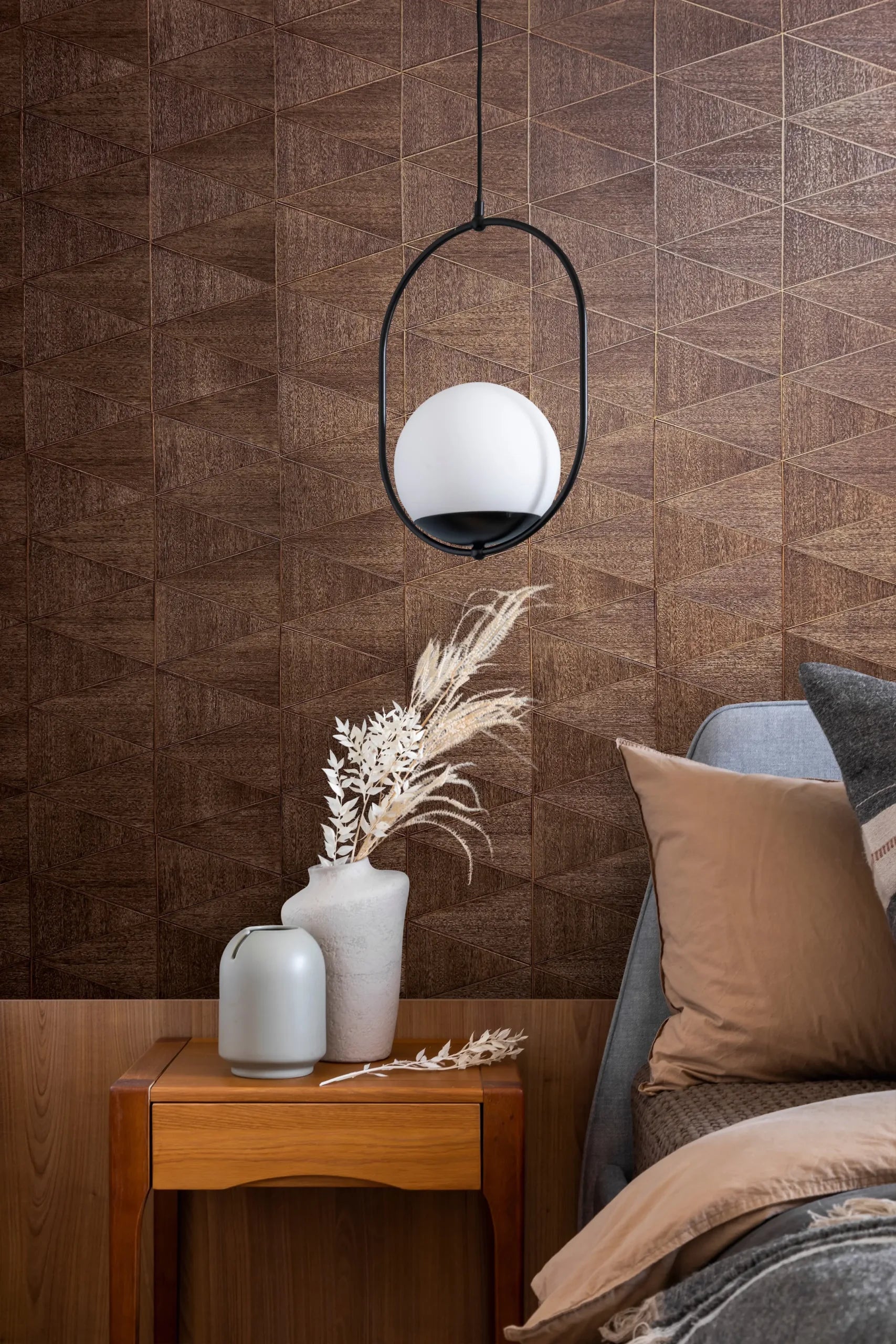 Marburg Travertino 33361 Brown geometric wallpaper with a woodgrain effect shown in a bedroom scene with a bedside table, two vases holding dried stems, a hanging pendant light, and part of a bed with pillows and a blanket.