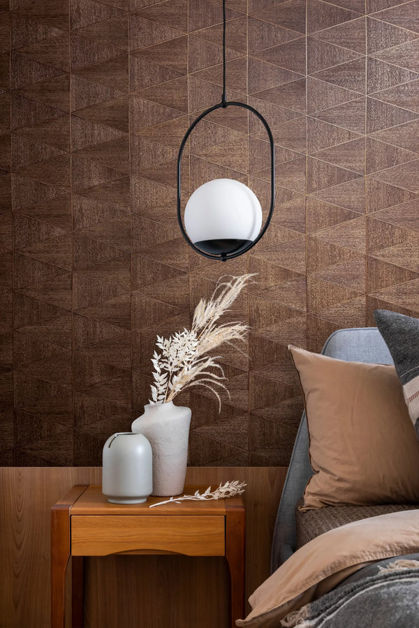 Marburg Travertino 33361 Brown geometric wallpaper with a woodgrain effect shown in a bedroom scene with a bedside table, two vases holding dried stems, a hanging pendant light, and part of a bed with pillows and a blanket.