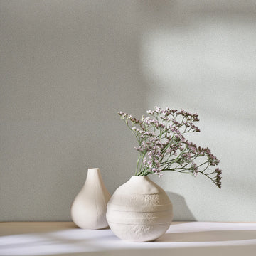 Marburg Travertino 33362 Light gray speckled wallpaper behind two white ceramic vases, one holding small purple flowers, with soft sunlight casting shadows.