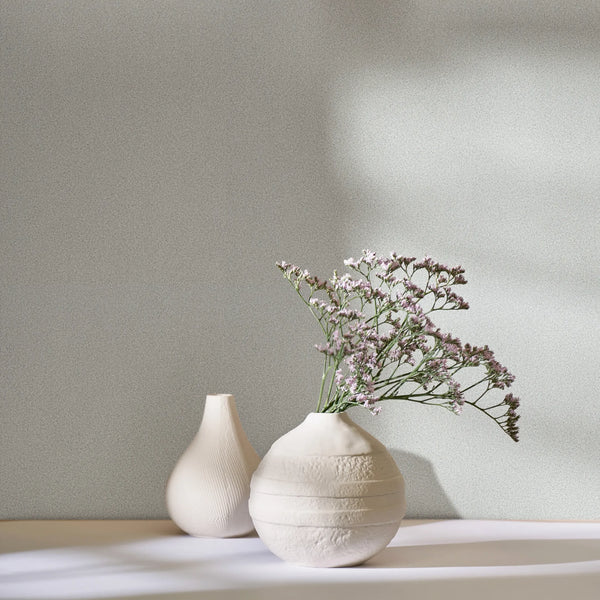 Marburg Travertino 33362 Light gray speckled wallpaper behind two white ceramic vases, one holding small purple flowers, with soft sunlight casting shadows.