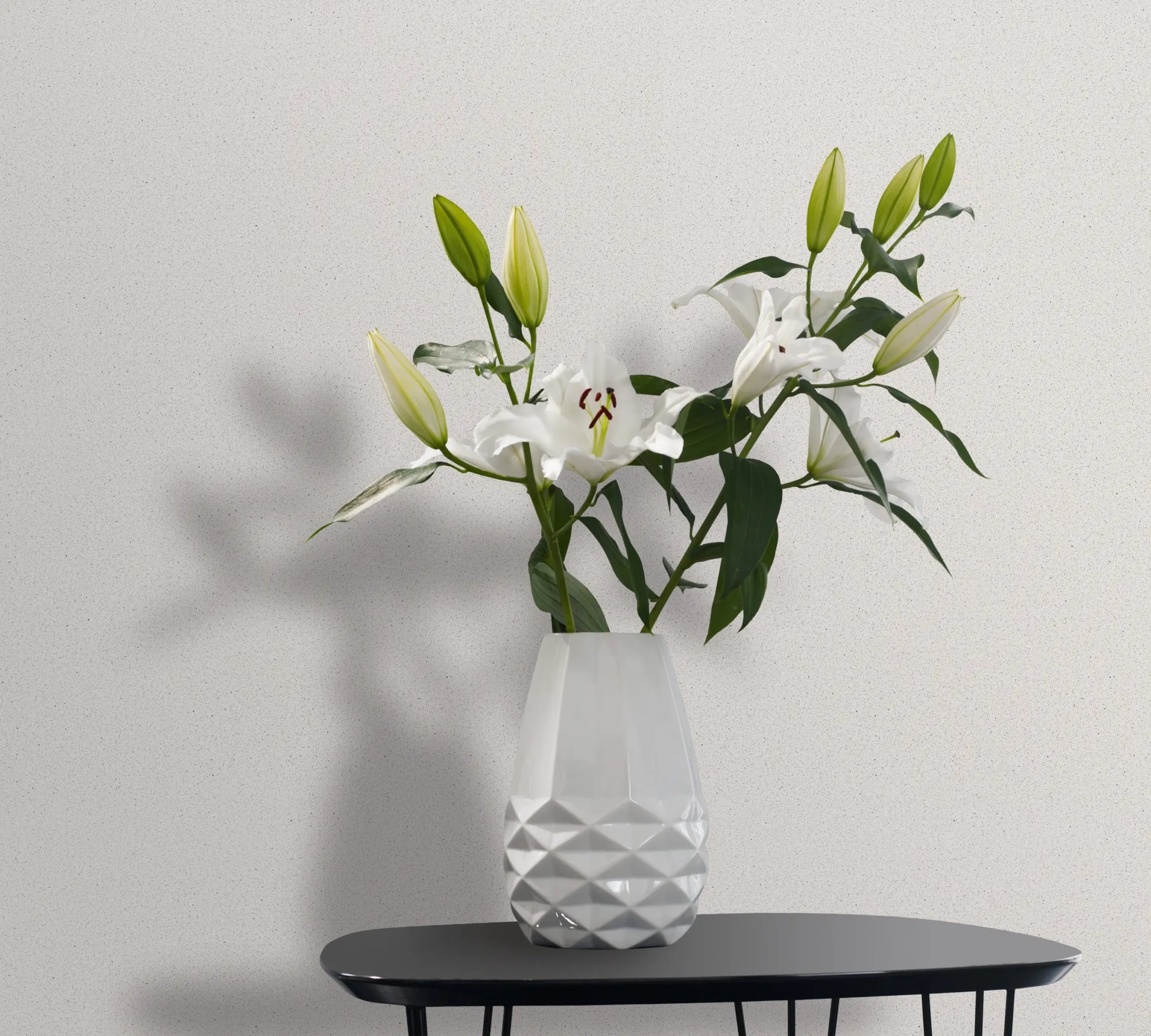 Marburg Travertino 33364 Light speckled white wallpaper behind a faceted white vase holding white lilies on a small black table.