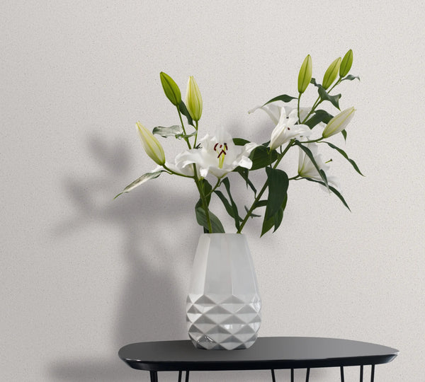 Marburg Travertino 33364 Light speckled white wallpaper behind a faceted white vase holding white lilies on a small black table.
