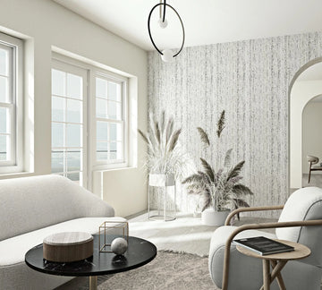 Marburg Travertino 33364 Bright living room featuring a white and gray vertically striated wallpaper on one wall, with large windows, a sofa, an armchair, a round black coffee table, indoor grasses in planters, and an arched doorway in view.