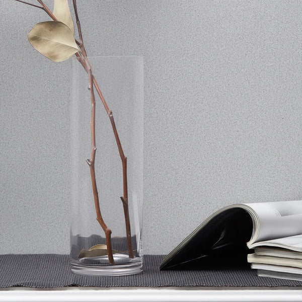 Marburg Travertino 33365 Light gray speckled wallpaper shown behind a clear glass cylinder vase with bare branches and an open magazine on a white molded console.