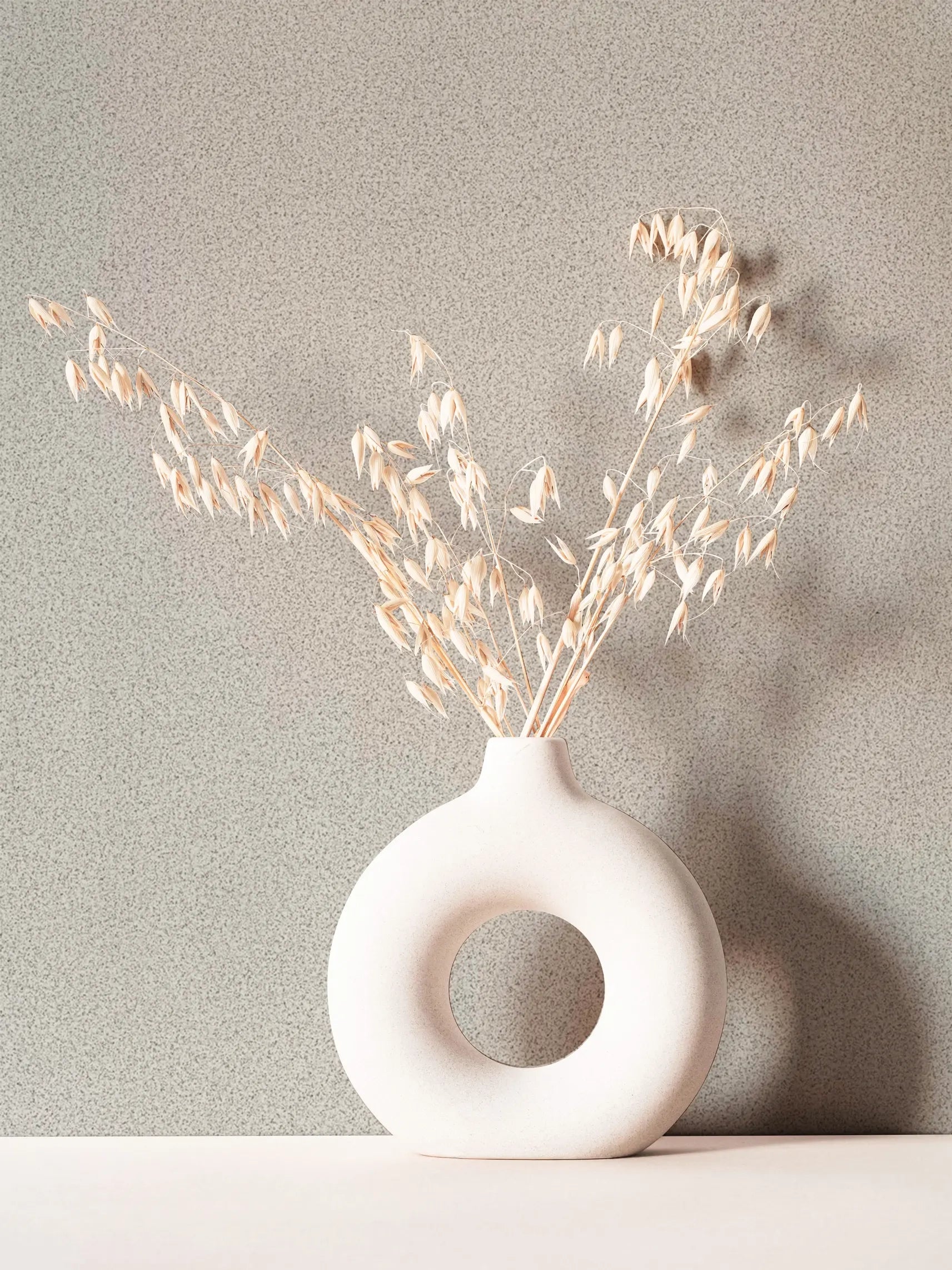 Marburg Travertino 33366 Light gray speckled wallpaper shown behind a round white ceramic ring vase holding dried stems on a pale tabletop.