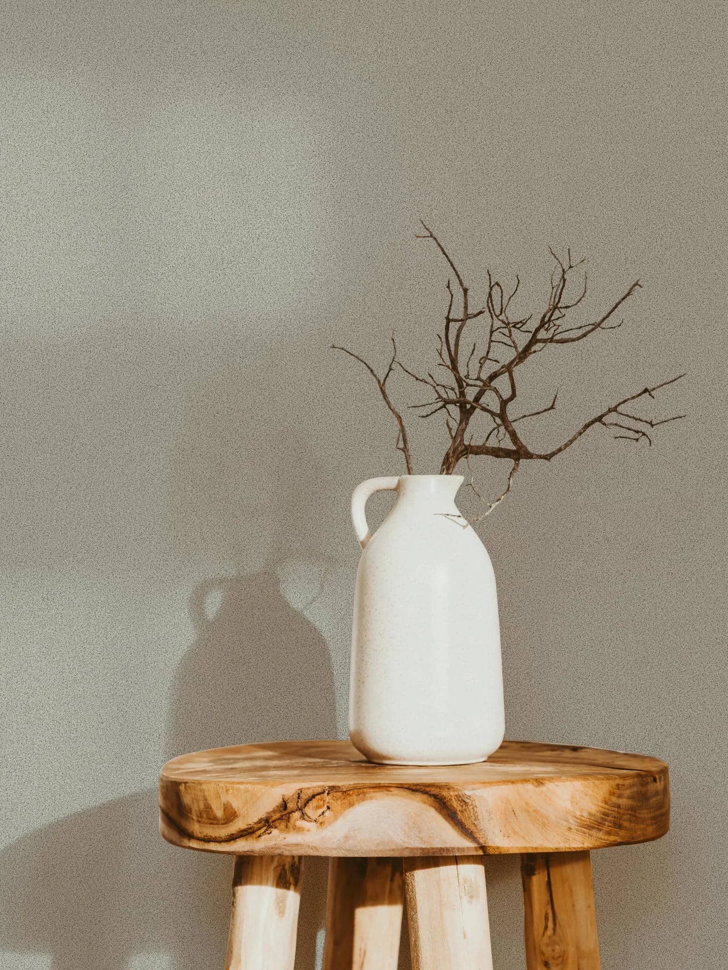 Marburg Travertino 33366 Subtle speckled gray wallpaper behind a white ceramic jug with bare branches on a wooden stool.
