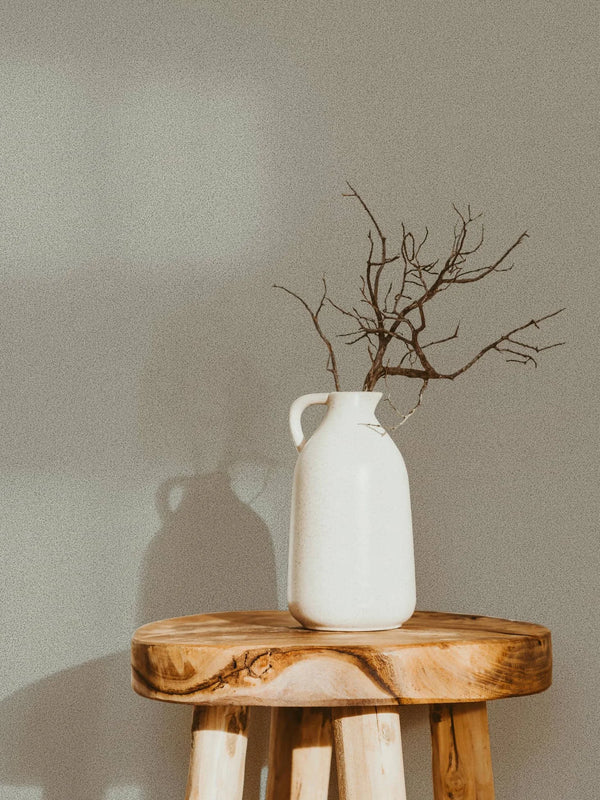 Marburg Travertino 33366 Subtle speckled gray wallpaper behind a white ceramic jug with bare branches on a wooden stool.