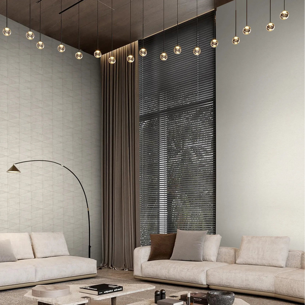 Marburg Travertino 33367 Wide living room scene showing light beige wallpaper; the left wall has a subtle small‑scale geometric triangle pattern and the adjacent wall is a plain textured surface. Two sofas, low coffee tables, an arc floor lamp, long curtains, window blinds, a rug, and.