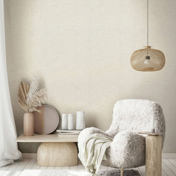 Marburg Travertino 33367 Beige textured wallpaper shown in a styled living room corner with a wicker pendant light, a fluffy white armchair, a small wooden side table with a candle, and a low wood bench holding a vase with dried leaves and grasses, a round tray, and candles; a sheer a.