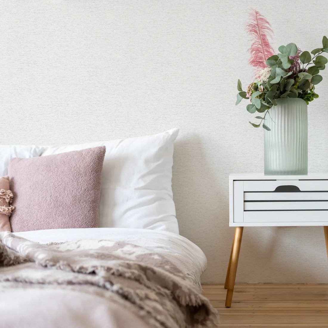 Marburg Travertino 33368 Light textured off-white wallpaper behind a styled bedroom scene with a bed, a small white bedside table topped with a ribbed glass vase and greenery, a floor vase with eucalyptus, and a white shelving unit.