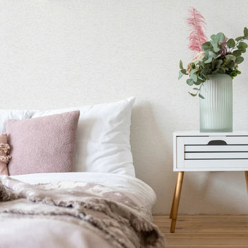 Marburg Travertino 33368 Light textured off-white wallpaper behind a styled bedroom scene with a bed, a small white bedside table topped with a ribbed glass vase and greenery, a floor vase with eucalyptus, and a white shelving unit.