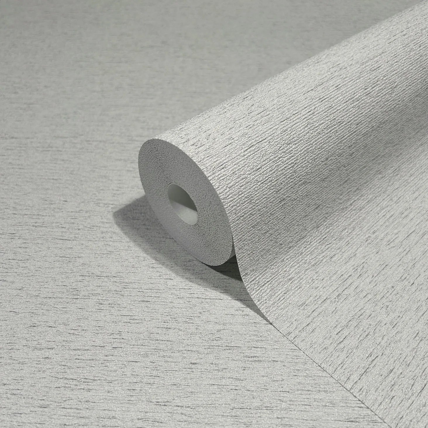 Marburg Travertino 33369 Close-up of a gray textured wallpaper roll resting on a matching unrolled sheet, showing a fine horizontal striated texture.