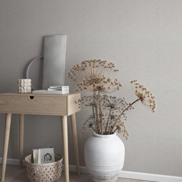Marburg Travertino 33369 Textured beige-gray wallpaper behind a light wood console table with books and a small ceramic piece, next to a large white vase holding dried seed heads; a woven basket sits on the floor by the baseboard.