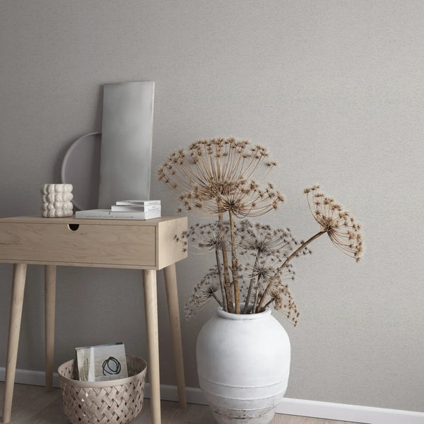 Marburg Travertino 33369 Textured beige-gray wallpaper behind a light wood console table with books and a small ceramic piece, next to a large white vase holding dried seed heads; a woven basket sits on the floor by the baseboard.
