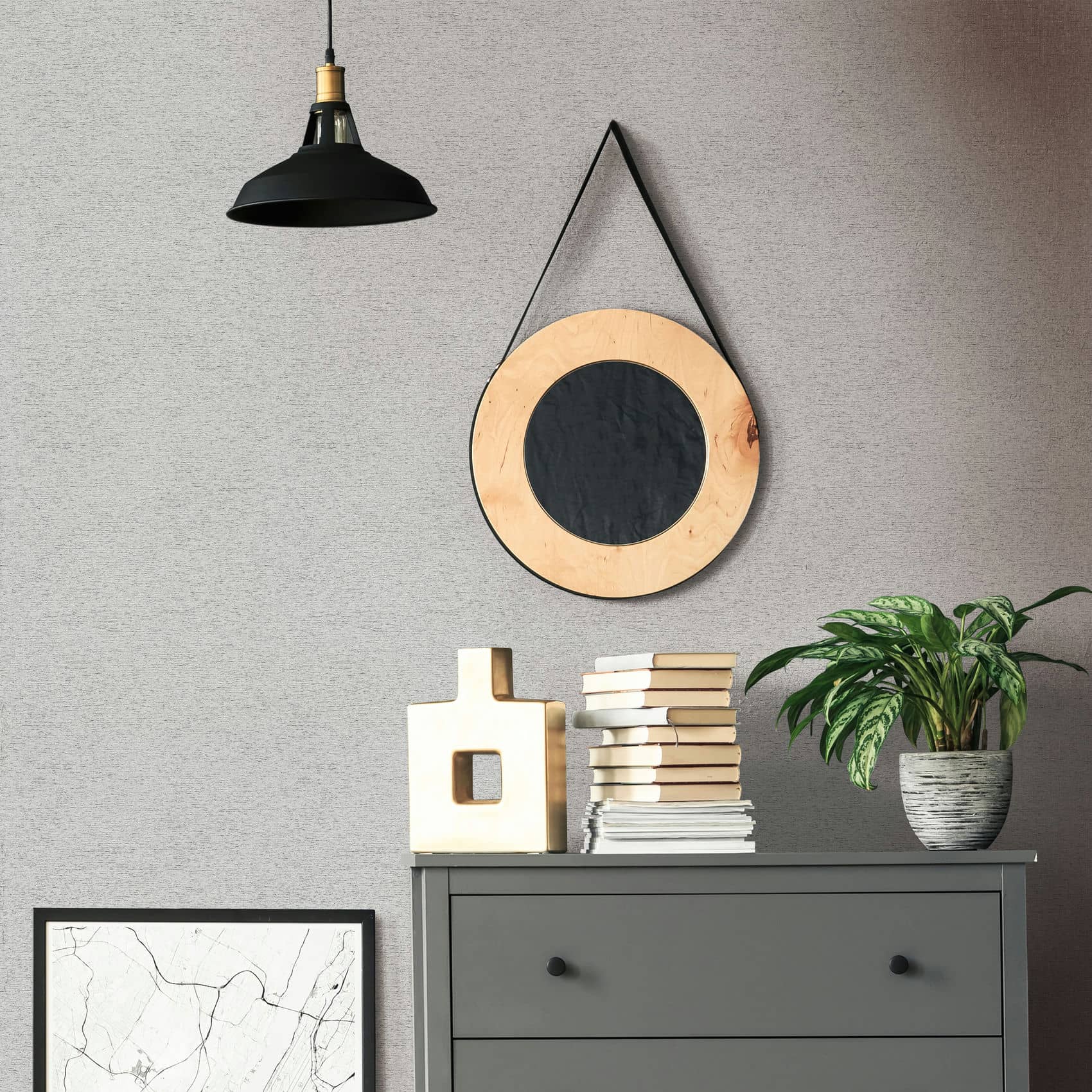Marburg Travertino 33370 Gray textured wallpaper behind a dark gray dresser with stacked books, a sculptural light-colored vase, and a potted plant; a round hanging mirror and a black pendant light above, with a framed print leaning against the wall.