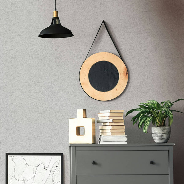 Marburg Travertino 33370 Gray textured wallpaper behind a dark gray dresser with stacked books, a sculptural light-colored vase, and a potted plant; a round hanging mirror and a black pendant light above, with a framed print leaning against the wall.