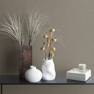 Marburg Travertino 33371 Brown textured wallpaper with fine horizontal lines behind a dark console styled with a tall rectangular vase of grasses, two white ceramic vases with dried stems, and a stack of magazines topped with a small candle.
