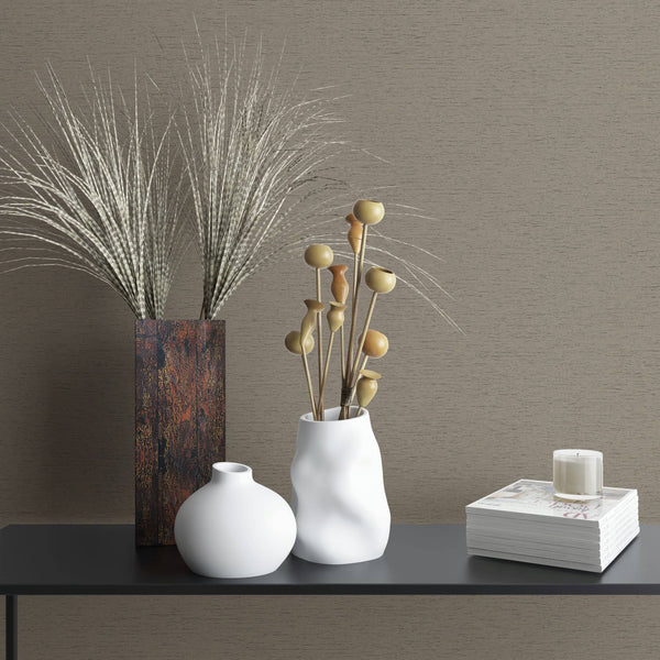 Marburg Travertino 33371 Brown textured wallpaper with fine horizontal lines behind a dark console styled with a tall rectangular vase of grasses, two white ceramic vases with dried stems, and a stack of magazines topped with a small candle.