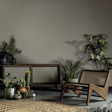 Marburg Travertino 33372 Warm brown textured wallpaper behind a cane-front sideboard, a wooden lounge chair, clustered potted plants, and a round woven rug.