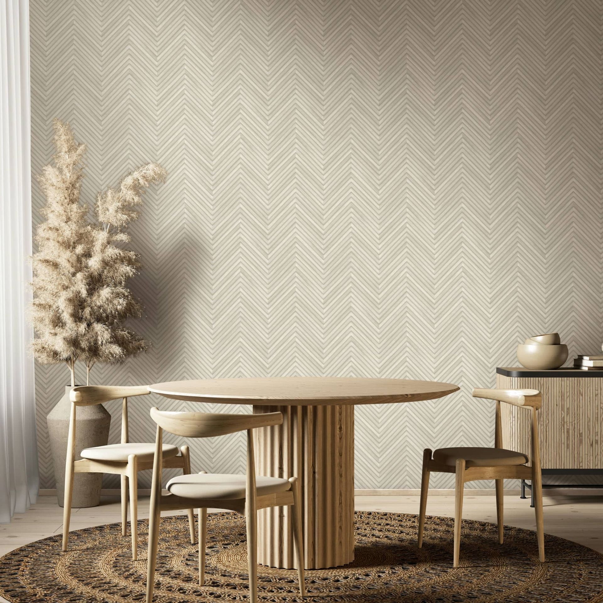 Marburg Travertino 33373 Beige herringbone wallpaper in a dining room setting with a round wooden table, three wooden chairs, a woven circular rug, a tall floor vase holding pampas grass, and a sideboard with decorative bowls.