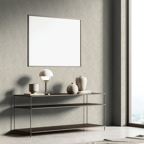Marburg Travertino 33374 Gray herringbone wallpaper in a styled room vignette with a slim console table, dome table lamp, ceramic vases, stacked books, and a large blank framed piece near a window.