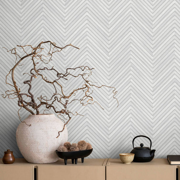 Marburg Travertino 33375 Light gray and white chevron stripe wallpaper behind a cabinet styled with a large ceramic vase holding twisting branches, a small jar, a black footed bowl with dried stems, a black teapot with a cup, and a closed book.