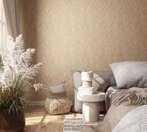 Marburg Travertino 33376 Beige chevron-patterned wallpaper shown in a bedroom setting with pillows and throw, a sculptural side table with books, a woven basket with cushions, a large pot of pampas grass, curtains, and a wood floor.