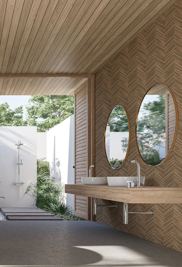 Marburg Travertino 33377 Brown chevron-pattern wallpaper behind a floating wood vanity with two vessel sinks and oval mirrors in a bright bathroom, with an open doorway showing a shower and greenery outside.