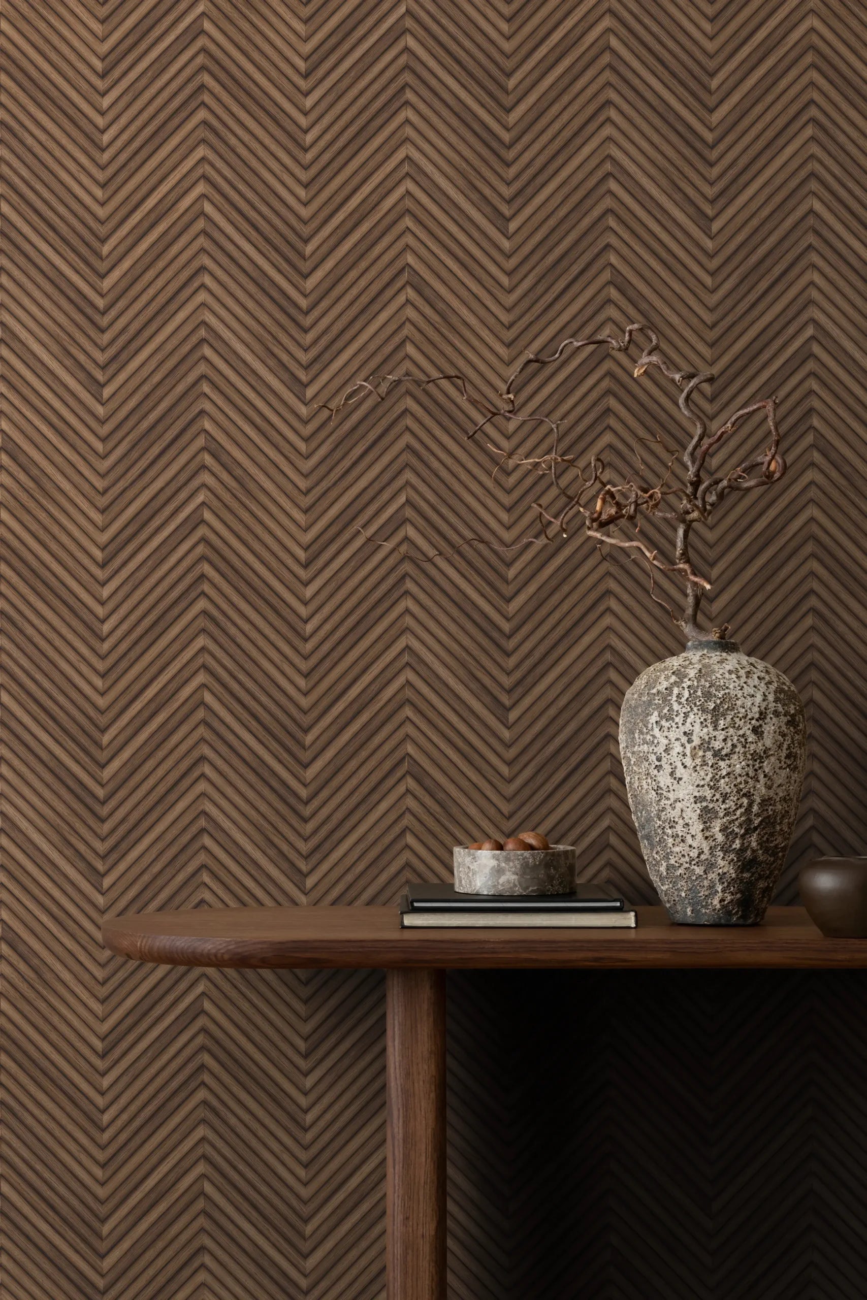 Marburg Travertino 33378 Brown chevron wallpaper behind a console table styled with a large textured vase holding a dried branch, a small bowl on books, and a small cup.