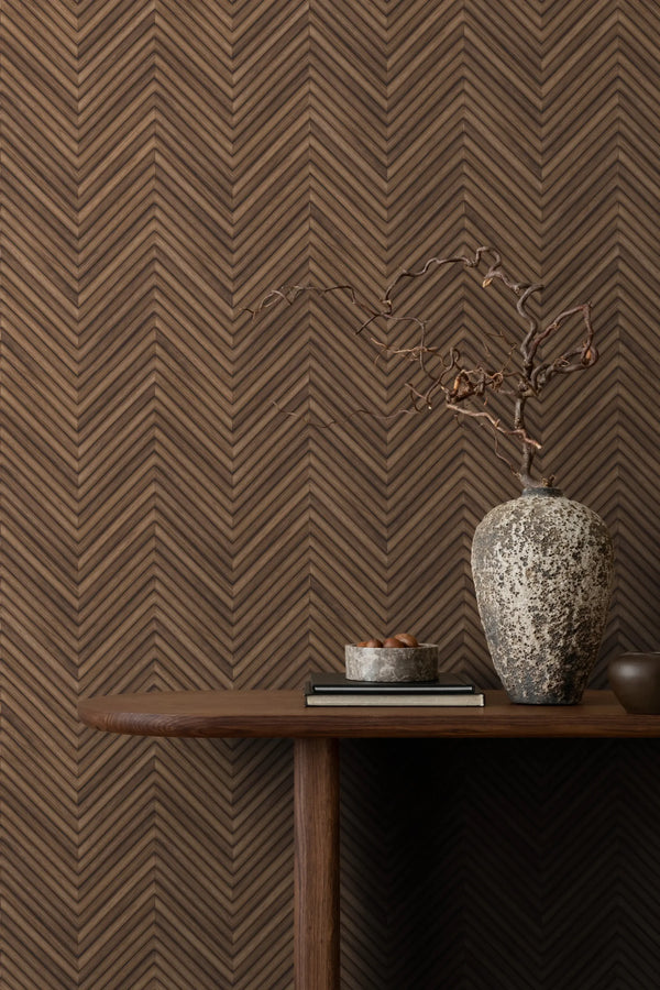 Marburg Travertino 33378 Brown chevron wallpaper behind a console table styled with a large textured vase holding a dried branch, a small bowl on books, and a small cup.