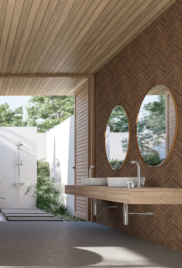 Marburg Travertino 33378 Brown chevron wallpaper behind a floating vanity with two oval mirrors and vessel sinks in a bright bathroom, with greenery and an outdoor shower visible.