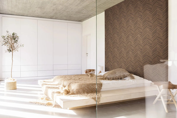 Marburg Travertino 33378 Bedroom with a brown herringbone wallpaper feature wall behind a low platform bed, with white built-in wardrobes, a potted tree, a small stool, and a table lamp.
