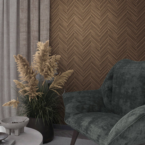 Marburg Travertino 33378 Brown chevron-pattern wallpaper behind a dark upholstered armchair, pampas grass in a black pot, grey curtains, and a small round table with a bowl.