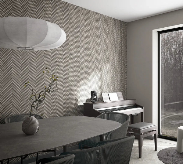 Marburg Travertino 33380 Gray chevron wallpaper on a feature wall in a dining area with a piano and bench, oval table with chairs, a pendant light, a small vase with branches, and a large window.