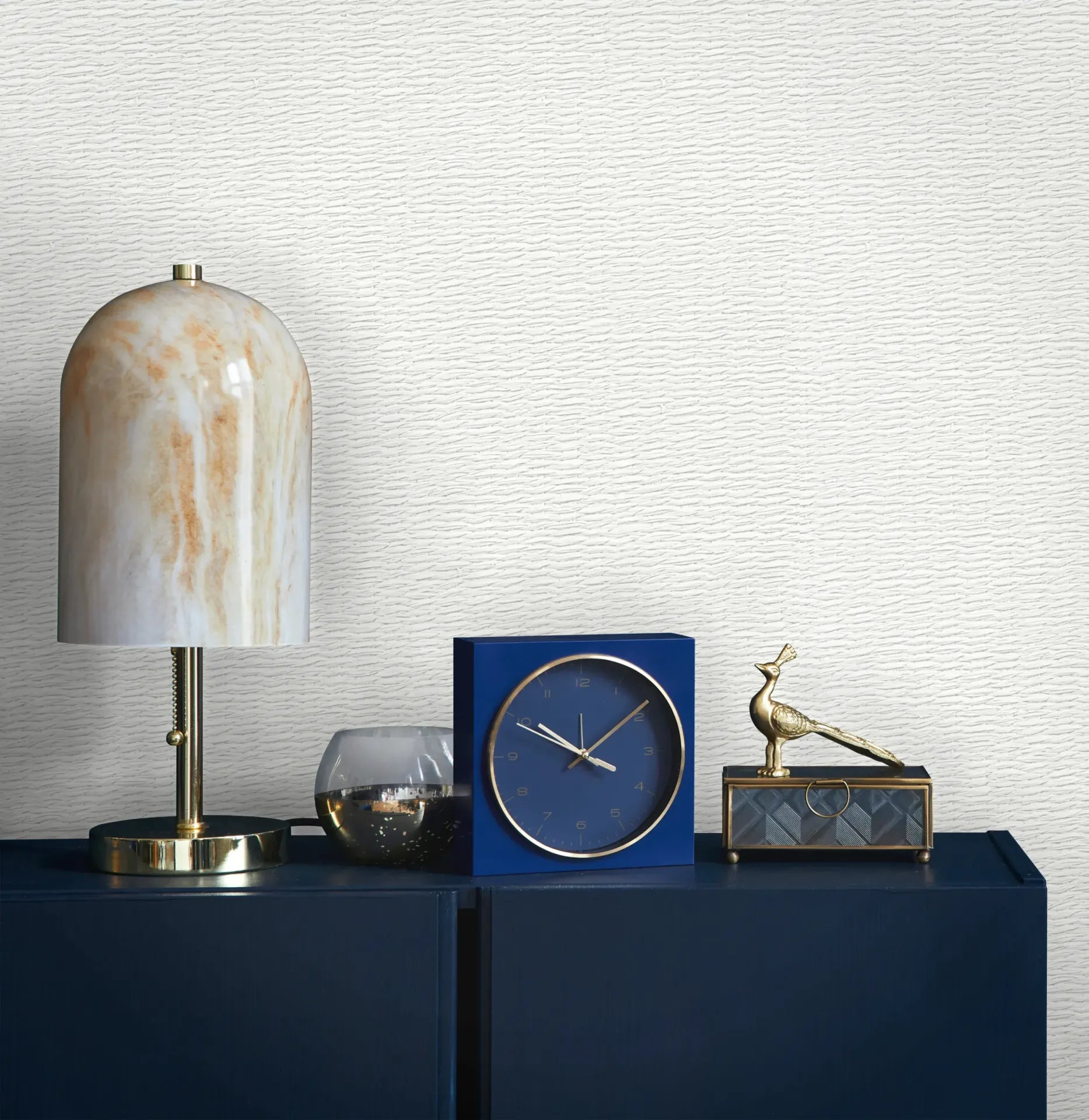 Marburg Travertino 33381 Light gray and white wallpaper with fine horizontal wavy lines behind a dark blue cabinet topped with a lamp with a marbled shade, a blue square clock, a small glass candle holder, and a metallic bird figurine on a decorative box.