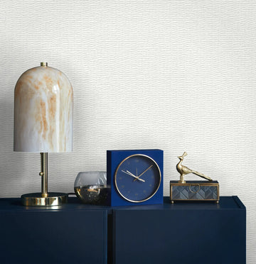 Marburg Travertino 33381 Light gray and white wallpaper with fine horizontal wavy lines behind a dark blue cabinet topped with a lamp with a marbled shade, a blue square clock, a small glass candle holder, and a metallic bird figurine on a decorative box.