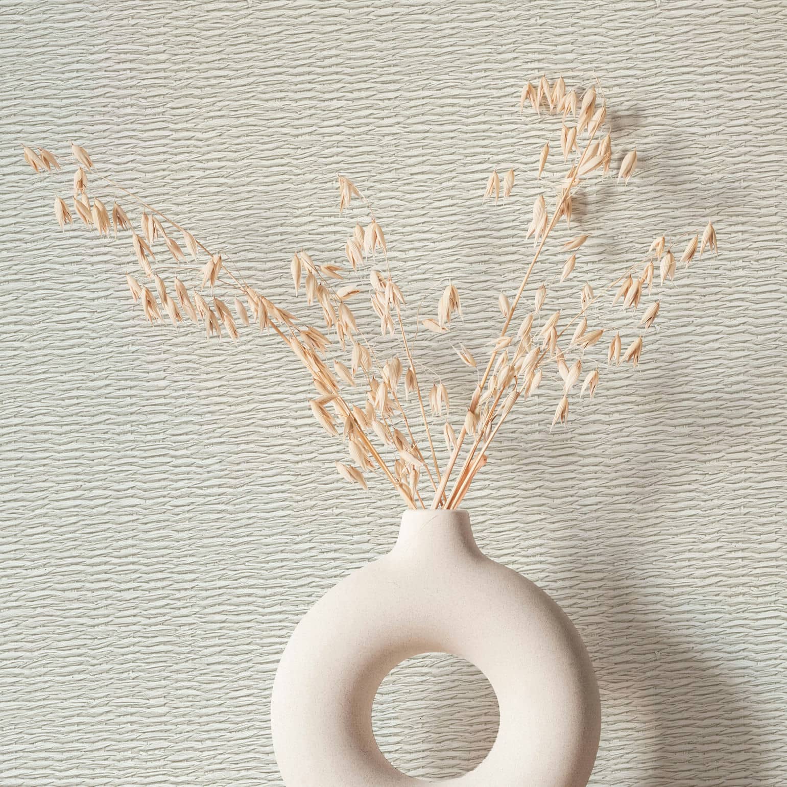 Marburg Travertino 33382 Close-up of light beige wallpaper with irregular horizontal ripple lines, styled with a sculptural ring-shaped vase holding dried stems.